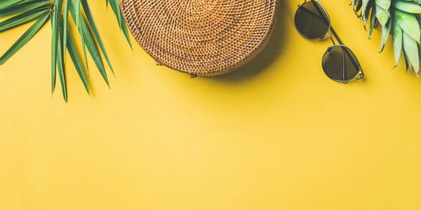 Beach essentials with a pineapple, sunglasses, palm leaves, and a woven bag on yellow.