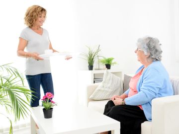 Woman brings tea to elderly lady in living room.