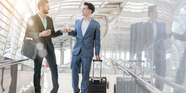 Two businessmen fist bump while walking in a modern transit area with luggage.