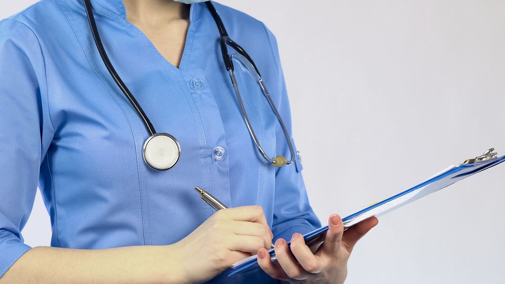 Healthcare professional in blue scrubs writing on a clipboard with a stethoscope around their neck.