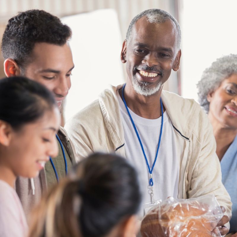 A cheerful mature male food bank volunteer stands in a warehouse with his coworkers and passes out bread to a young woman and her younger sister.