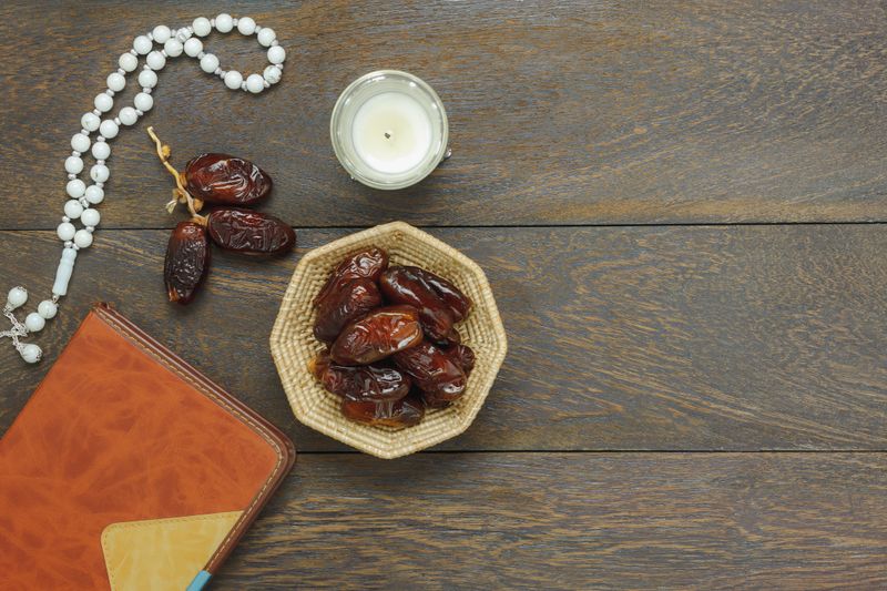 Table top view aerial image of decoration Ramadan Kareem holiday background.Flat lay date in wood basket with white prayer beads & lighting and holy book.The blackboard space on brown wood at office desk.