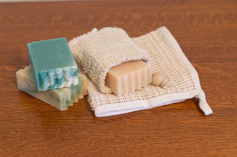 Bars of goats milk soap with one bar in a ramie soap caddy. A ramie washcloth is under the soap. A medium brown wood table is shown.