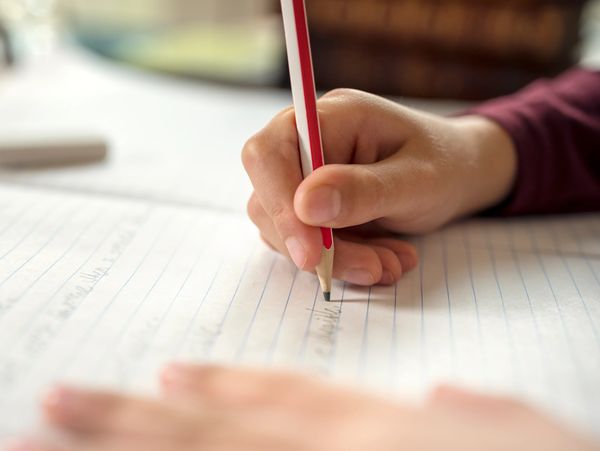 Close-up of a child’s hand writing with a pencil on lined paper in a notebook.