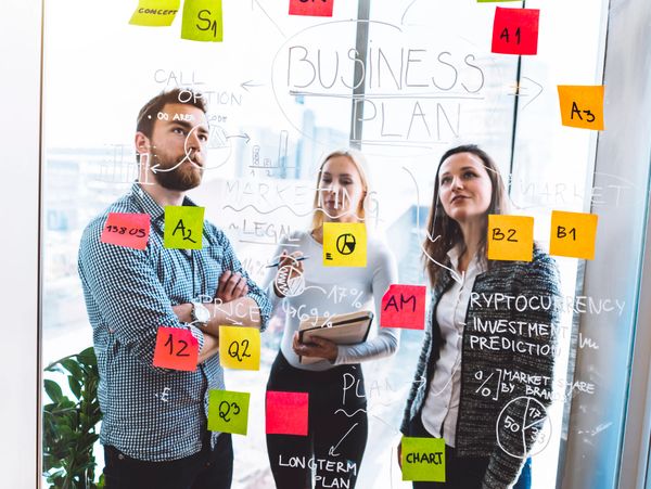 Three professionals discussing a business plan with colorful sticky notes on glass.