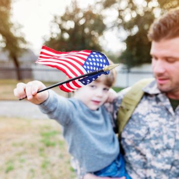 A soldier holding a child waving a small American flag outdoors.