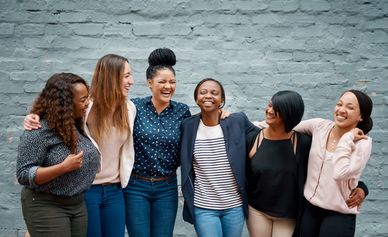 Group of six women happily standing together against a gray wall.