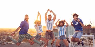 Group of friends joyfully jumping on a rooftop at sunset, wearing soccer jerseys.