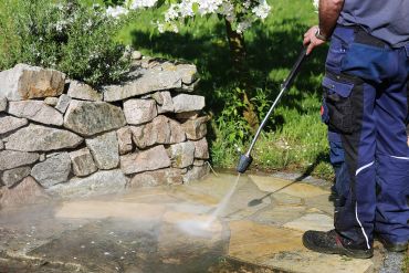 Person pressure washing stone pavement near a stone wall outdoors.