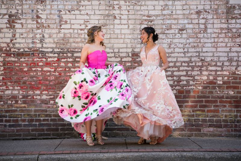 Two teenage girls wearing long party dresses dance together by a weathered brick wall outdoors on a beautiful spring day, Indiana, USA