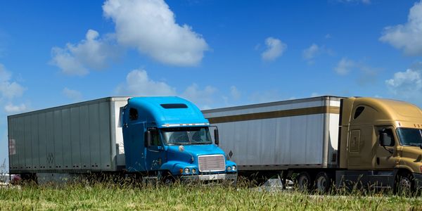 Two semi-trucks parked on grassy land under a bright blue sky.
