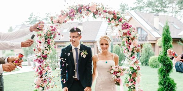 Newlywed couple joyfully walking under a floral arch as guests shower them with confetti.