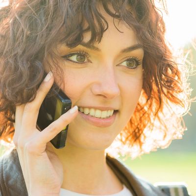A smiling woman with curly brown hair is talking on a black smartphone outdoors in bright sunlight.