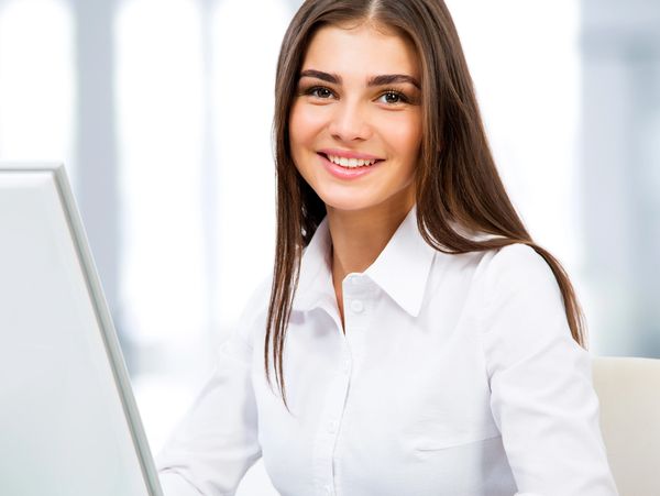 Smiling woman in a white shirt working at a laptop in a bright office.