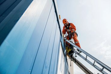 Worker in orange safety gear climbing a ladder on the side of a blue building.