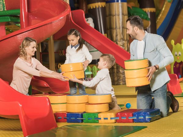Family playing together with large foam blocks near a red slide in a colorful indoor playground.