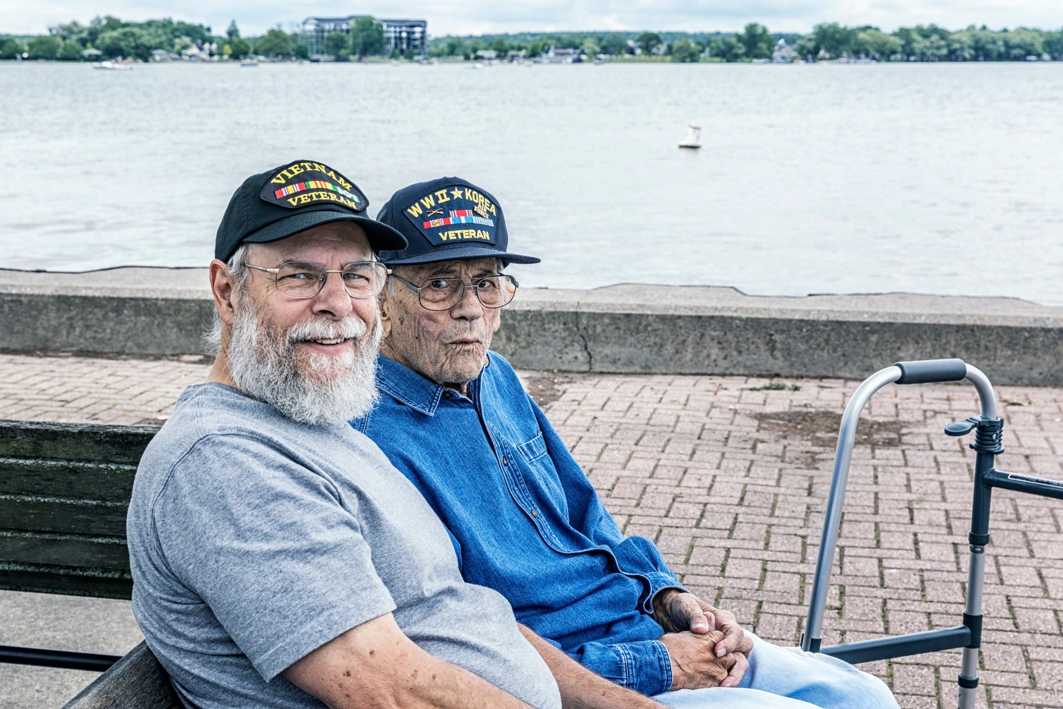Two elderly veterans sitting by a waterfront, wearing hats honoring their service.