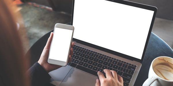 Person using a laptop and smartphone with blank white screens beside a cup of coffee.