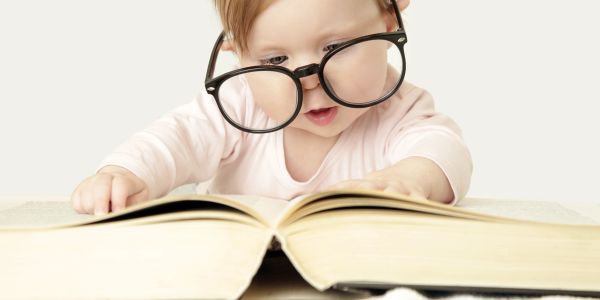 Baby wearing oversized glasses reads a large book with curiosity.