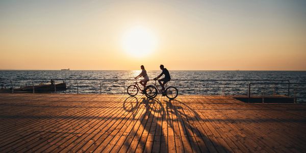 Two people cycling on a pier during sunset with long shadows on wooden planks.