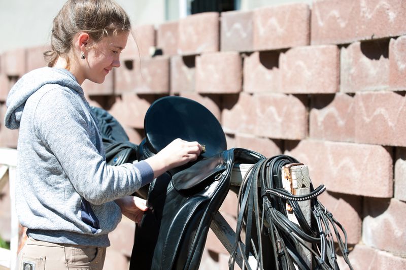Pretty teenage girl equestrian cleans black Leather Horse Saddle and equipment at farm on bright sunny day. Horizontal outdoors summertime image.