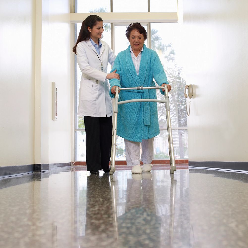 Nurse assisting elderly woman walking with a walker in a hospital corridor.