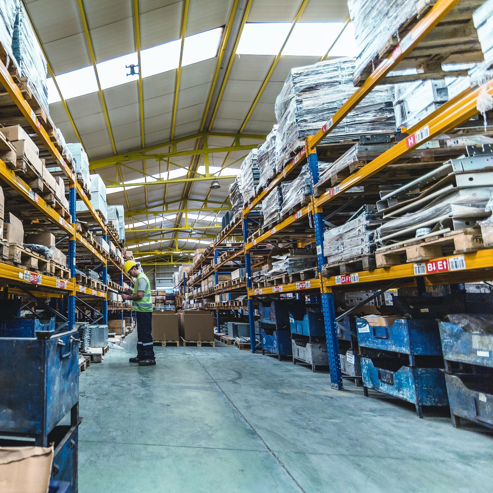 Warehouse worker inspecting inventory among tall shelves filled with boxes and pallets.