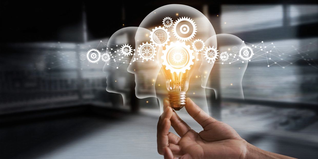 Hand holding a glowing light bulb with heads and gears representing ideas and innovation.