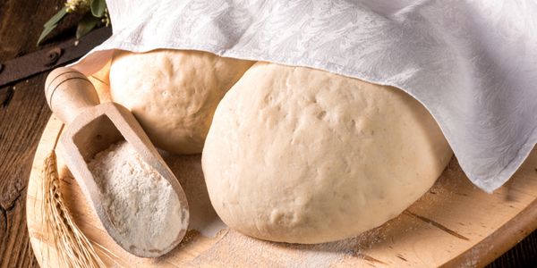 Two dough balls covered partially with a cloth on a wooden board with flour scoop.