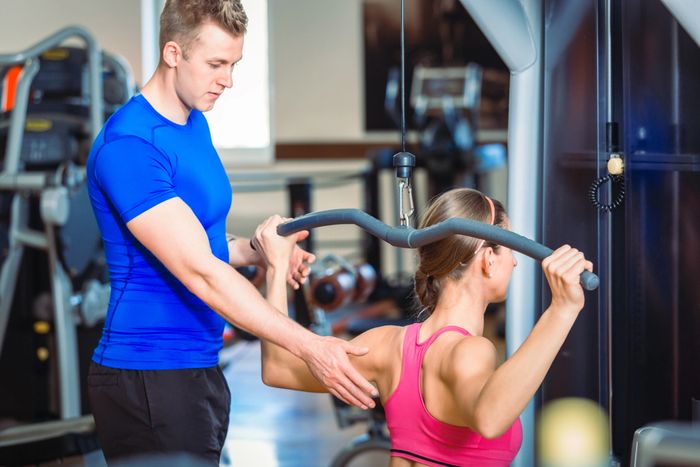 Personal trainer assisting woman with lat pulldown exercise in gym.