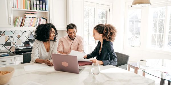 Three people discussing documents at a bright kitchen table with a laptop.