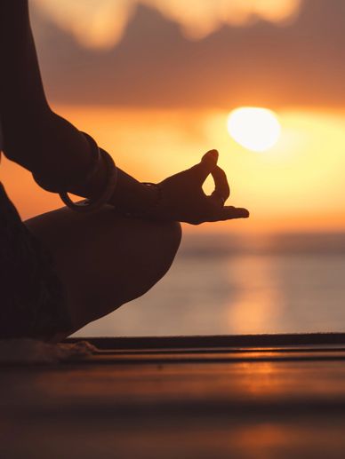 Person meditating at sunset near water, hand in mudra gesture.