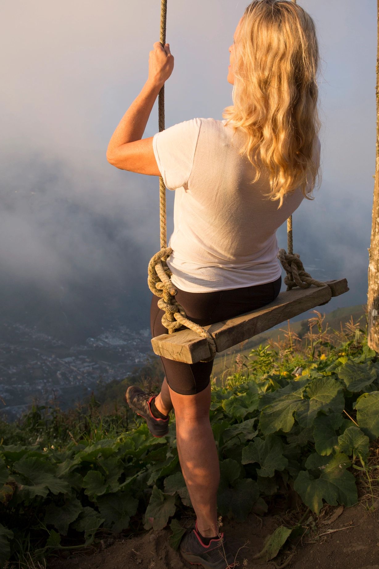 Woman with blonde hair on a wooden swing overlooking foggy mountains.