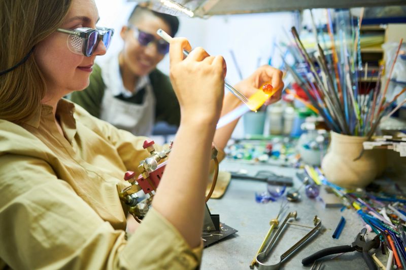 Side view portrait of young female artist melting glass by gas torch while making beads for beautiful handmade jewelry in glassworking studio