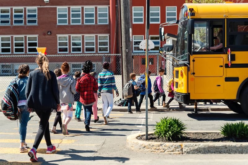Large group of multi-ethnic elementary age school kids crossing the street in line to get on the school bus. School in the background. Most of them are carrying backpack and lunch box. Horizontal full length outdoors shot on a bright sunny day with copy space.