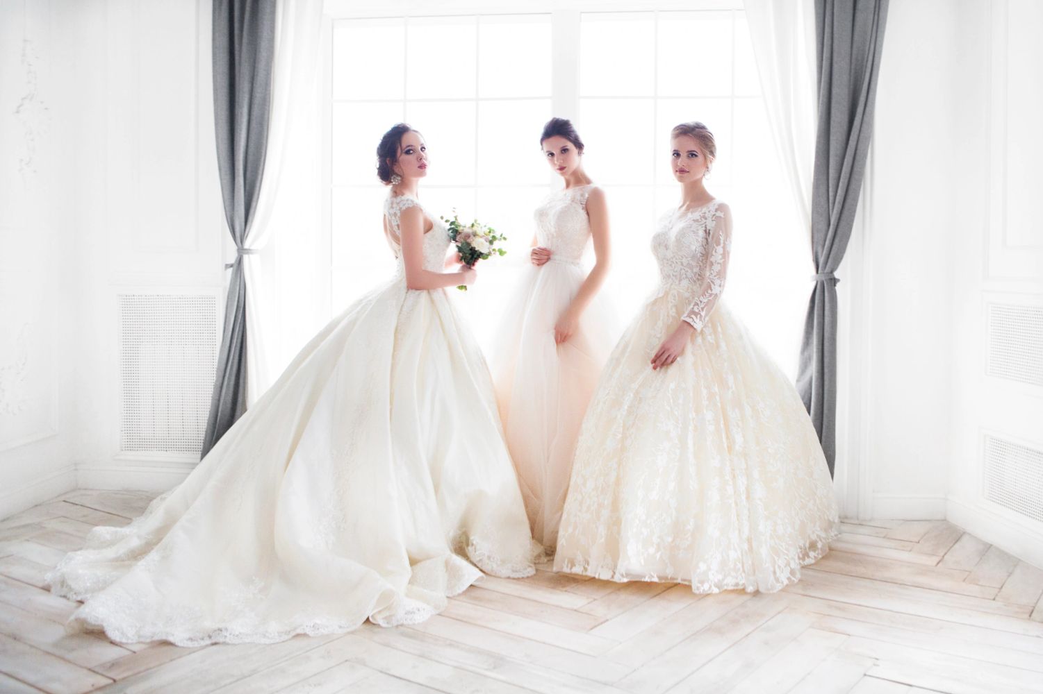 Three women in elegant wedding dresses posing by a window.