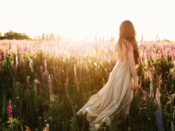 Woman in flowing dress walks through a sunlit flower field at sunset.