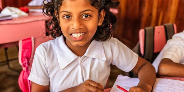Smiling schoolgirl in white uniform sitting at desk with workbook and pencil.
