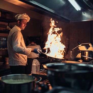 Chef flambéing food in a professional kitchen with large flames.