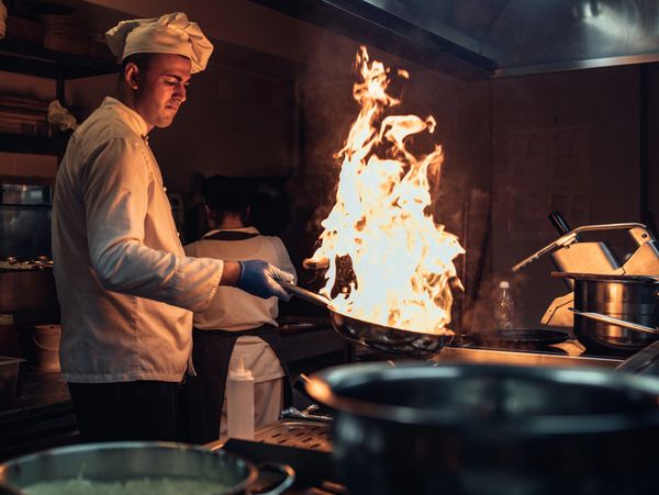 Chef flambéing food in a busy kitchen with intense flames.