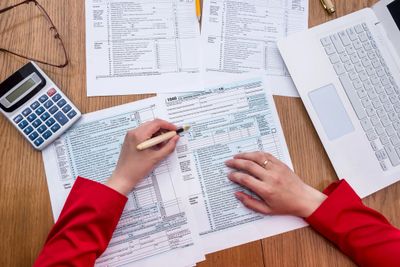 Person filling out a tax form with calculator and laptop nearby.