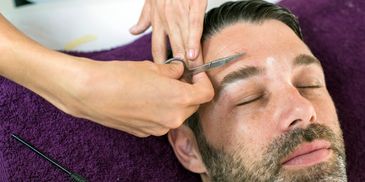 Man getting his eyebrows trimmed with scissors while lying on a purple towel.