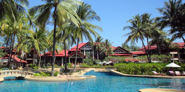 A tropical resort pool surrounded by palm trees and red-roofed buildings under a clear blue sky.