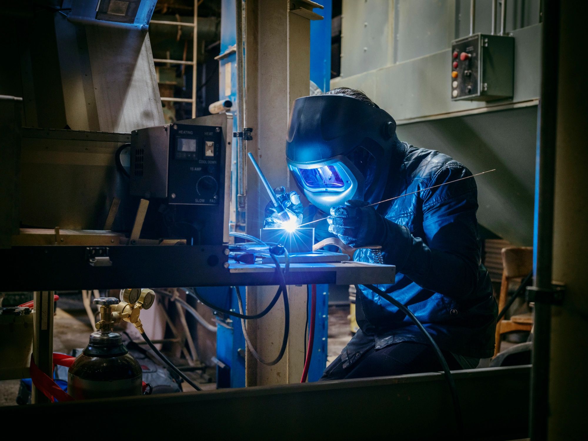 A welder in protective gear working on metal with a bright blue welding spark.