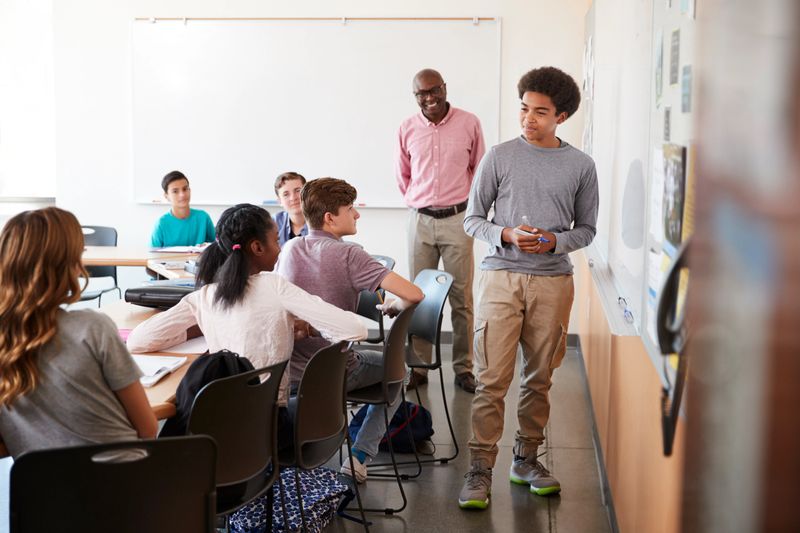 View Through Doorway Of High School Pupil Writing On Whiteboard In Class