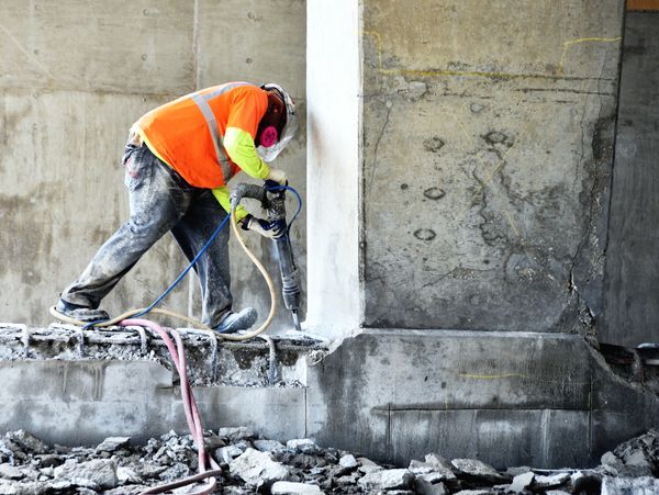 Construction worker using jackhammer on a concrete surface wearing safety gear.