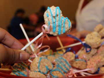 Hand holding a heart-shaped rice crispy treat with blue icing and a pink ribbon.