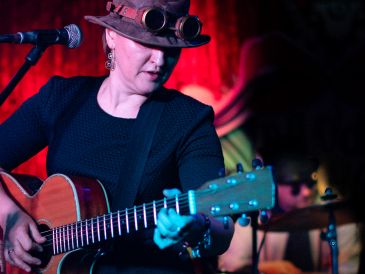 Musician playing guitar on stage with red lighting and steampunk hat.