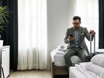 The image shows a man, dressed in a grey suit and patterned shirt sitting on the edge of a hotel bed