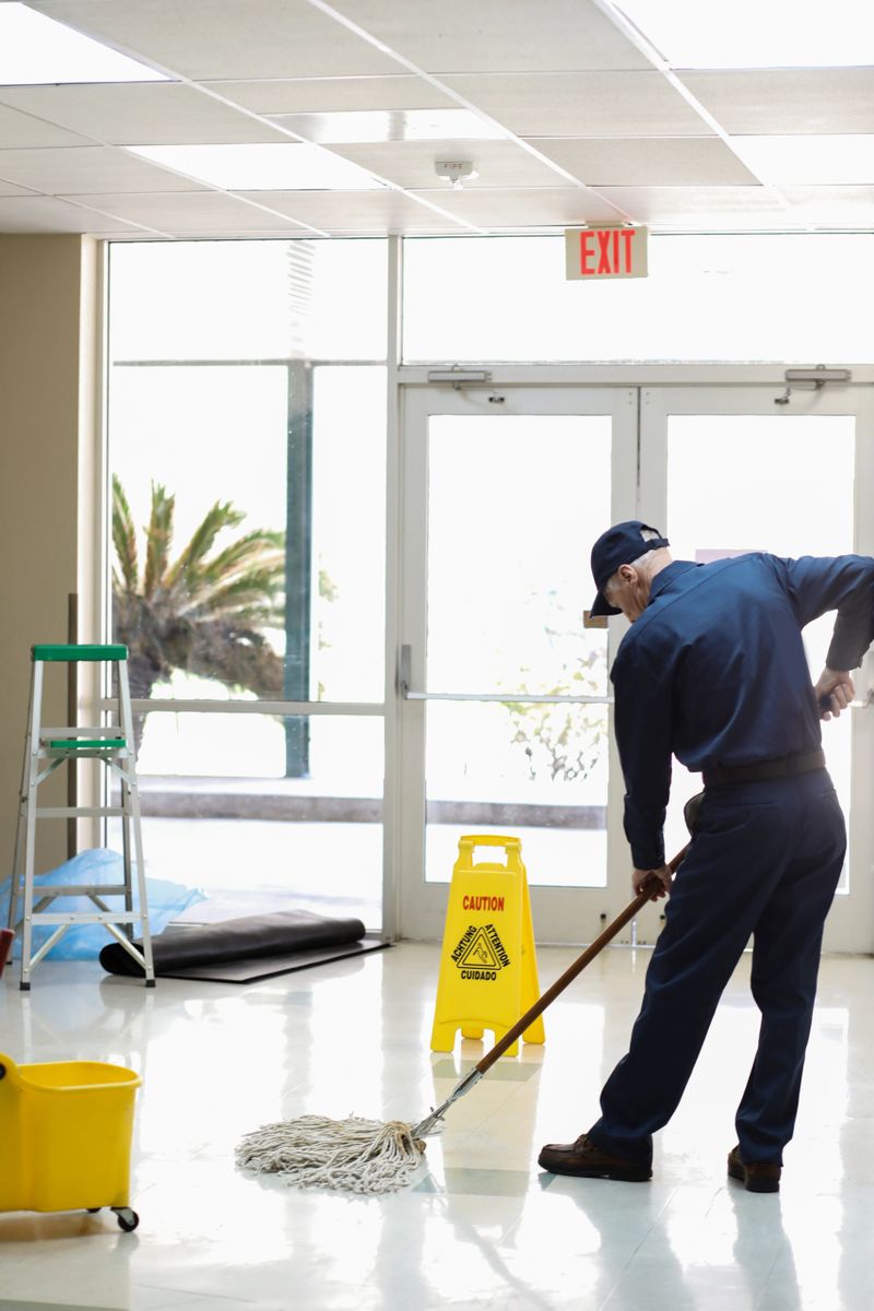 Senior adult man working as a janitor in an office building, school or church.  He wears a uniform as he mops the entry floor.  Mop bucket and safety sign are also pictured.